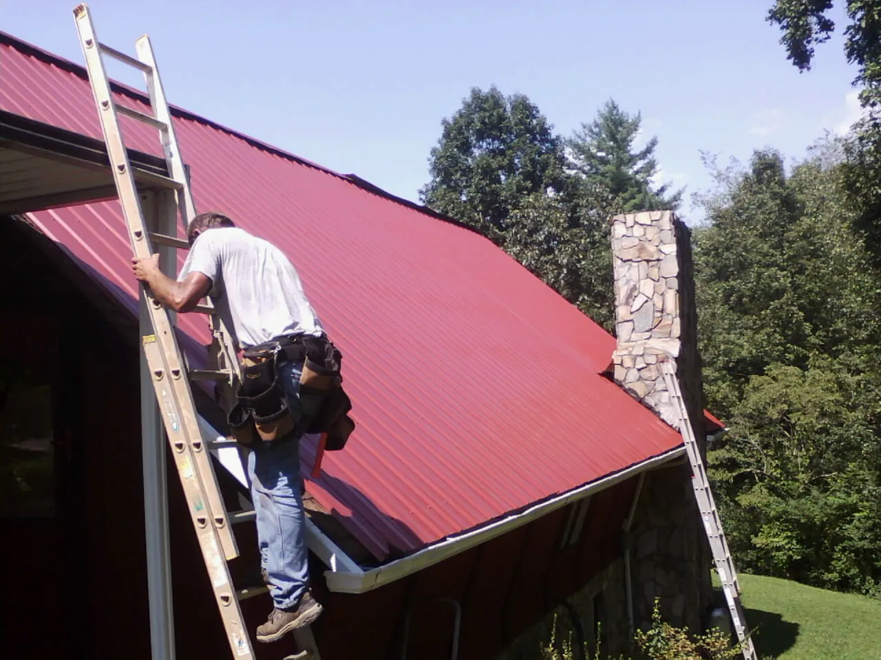 Roofer on a ladder beside a red metal roof and stone chimney — Soffit & Fascia Repair by Roofing Brothers in Herrick