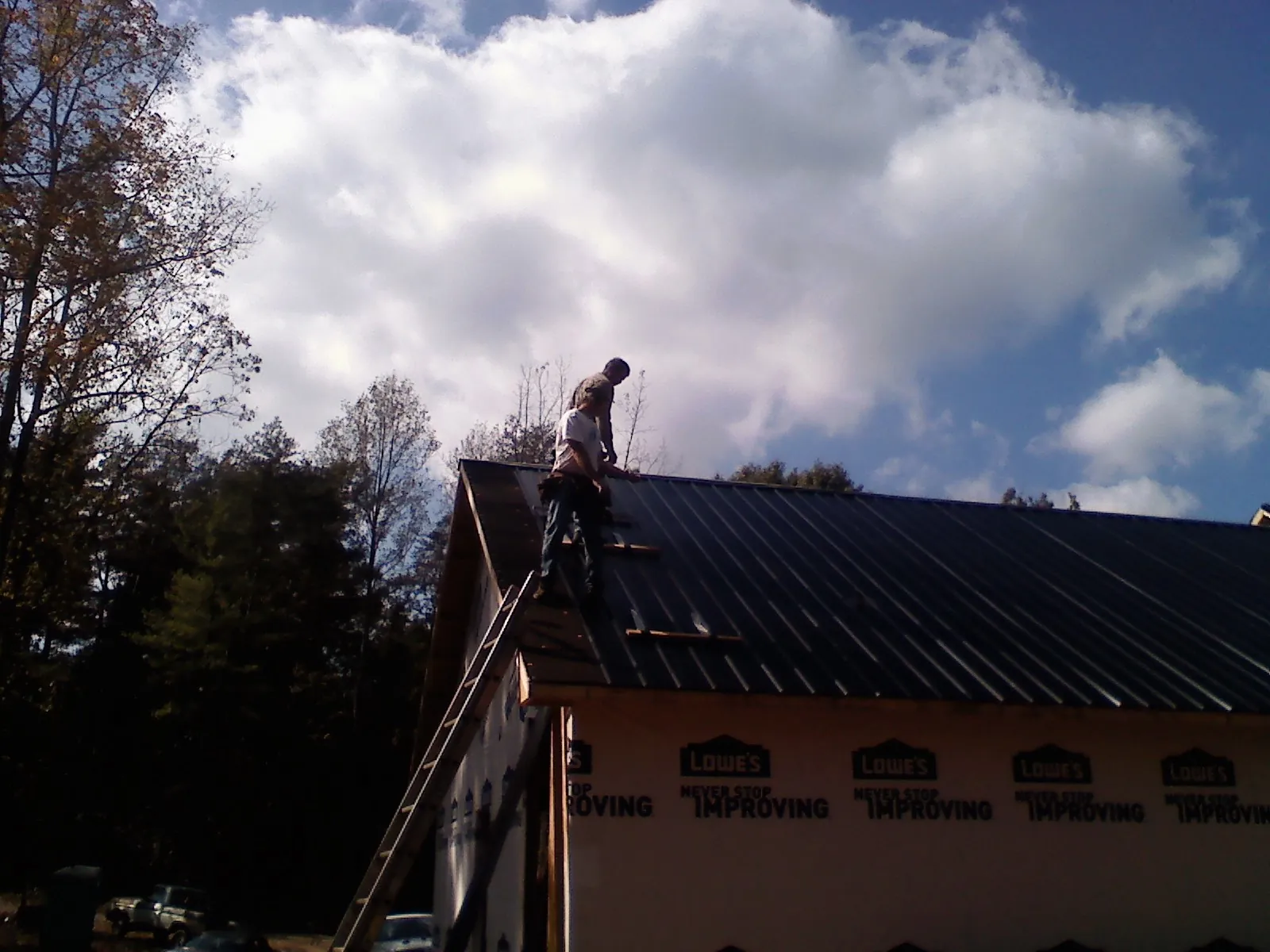 Roofer fastening dark metal panels high against a cloudy sky, from a Roof Inspection job in Herrick